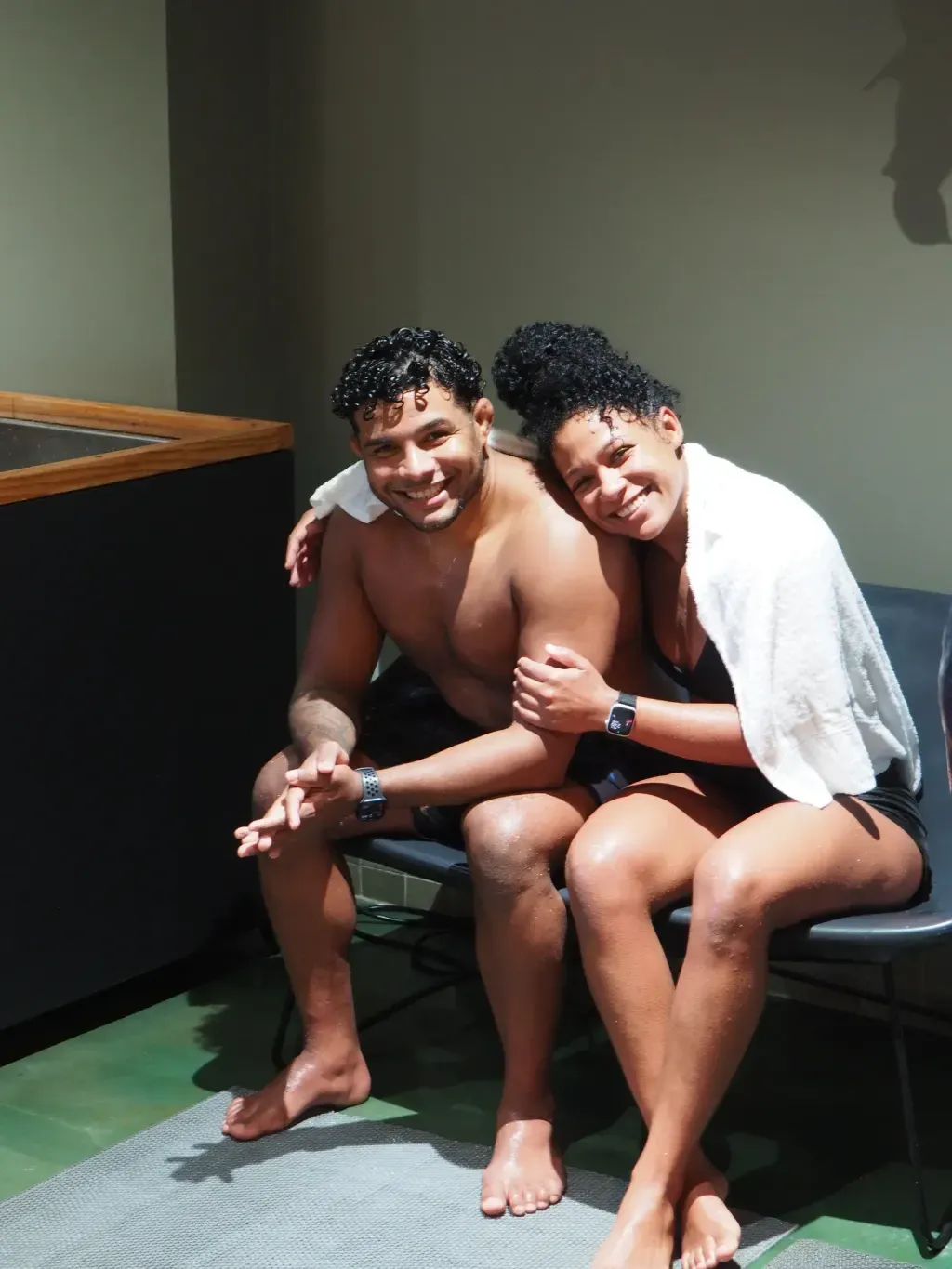 Couple smiling together on the cold-plunge room bench, wrapped in a towel, at Sauna Culture Charlotte