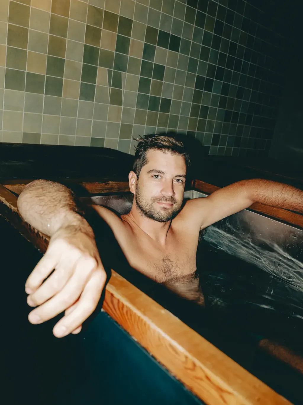 Guest chest-deep in an individual wood-rimmed cold plunge against the tiled mural at Sauna Culture Charlotte