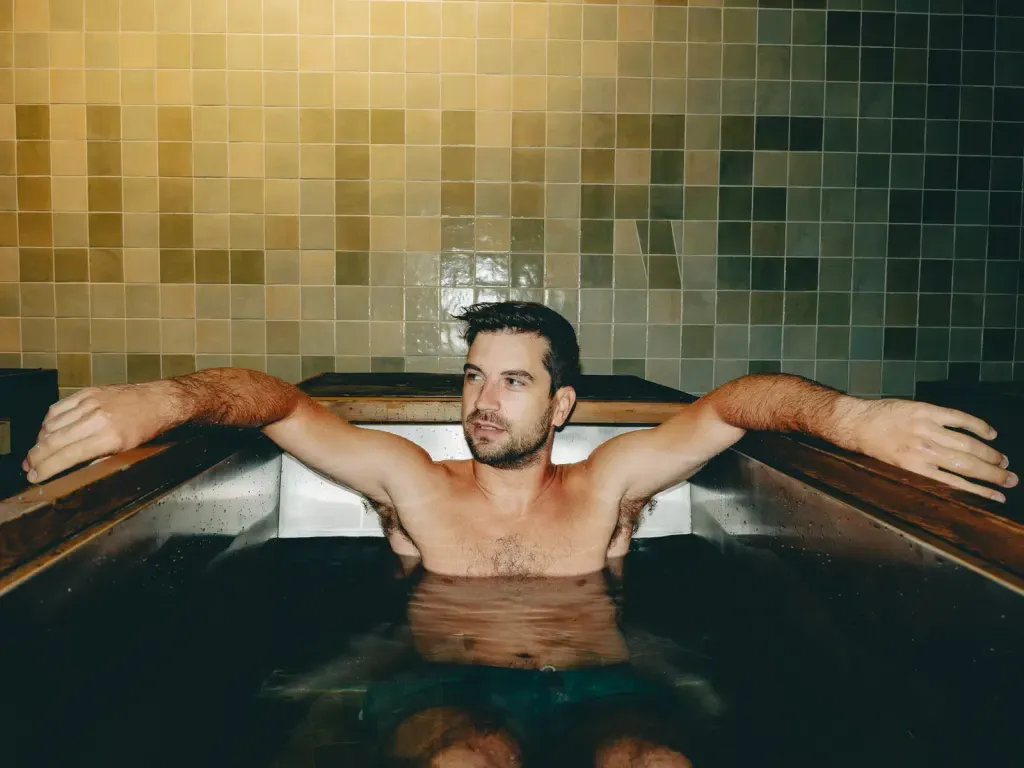 Guest with arms along the rim of an individual cold plunge in front of the Sauna Culture tile mural in Cotswold, Charlotte