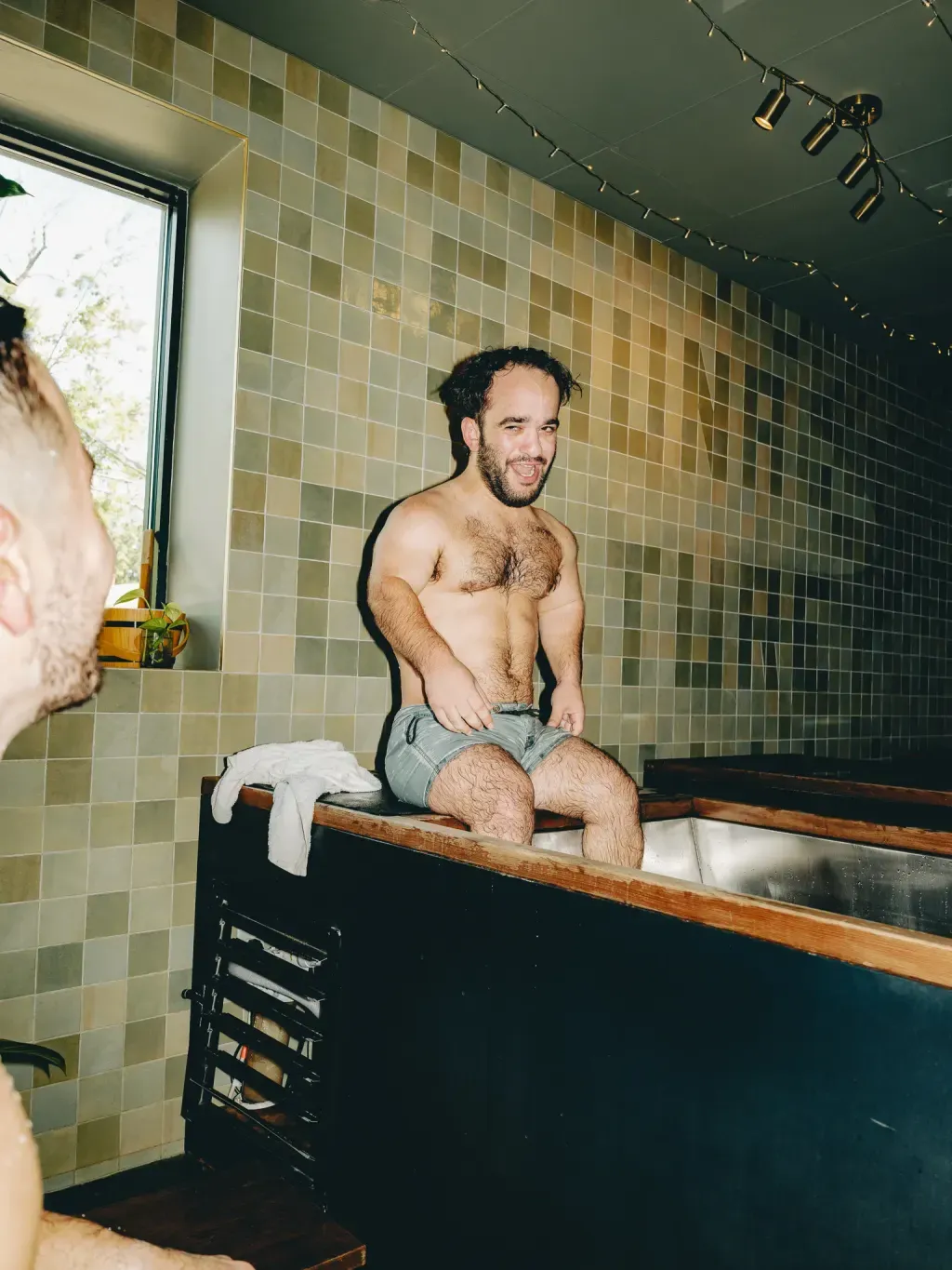 Guest laughing on the rim of an individual cold plunge in front of the Sauna Culture tile mural in Charlotte