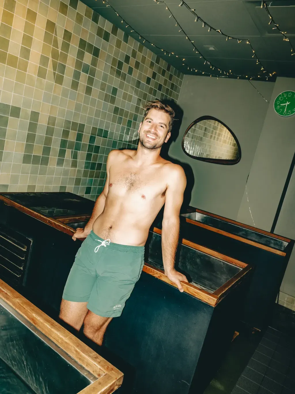 Guest smiling against an individual cold plunge beside the studio clock at Sauna Culture Charlotte