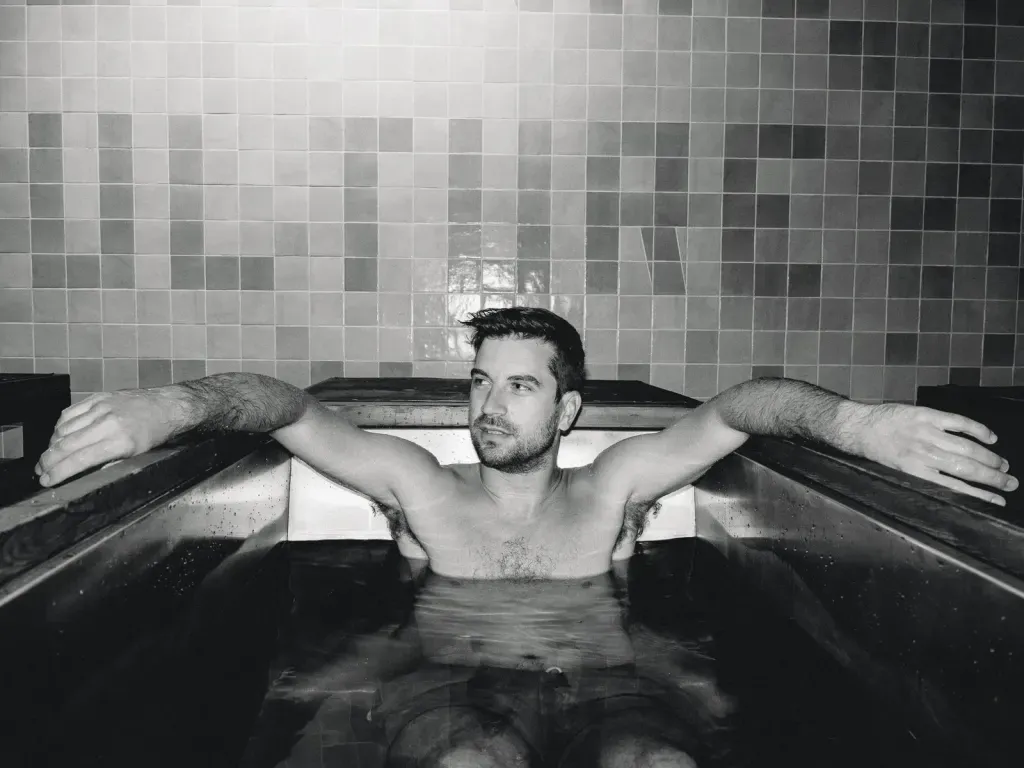 Black-and-white landscape of a guest in an individual cold plunge in front of the CULTURE tile mural at Sauna Culture Charlotte