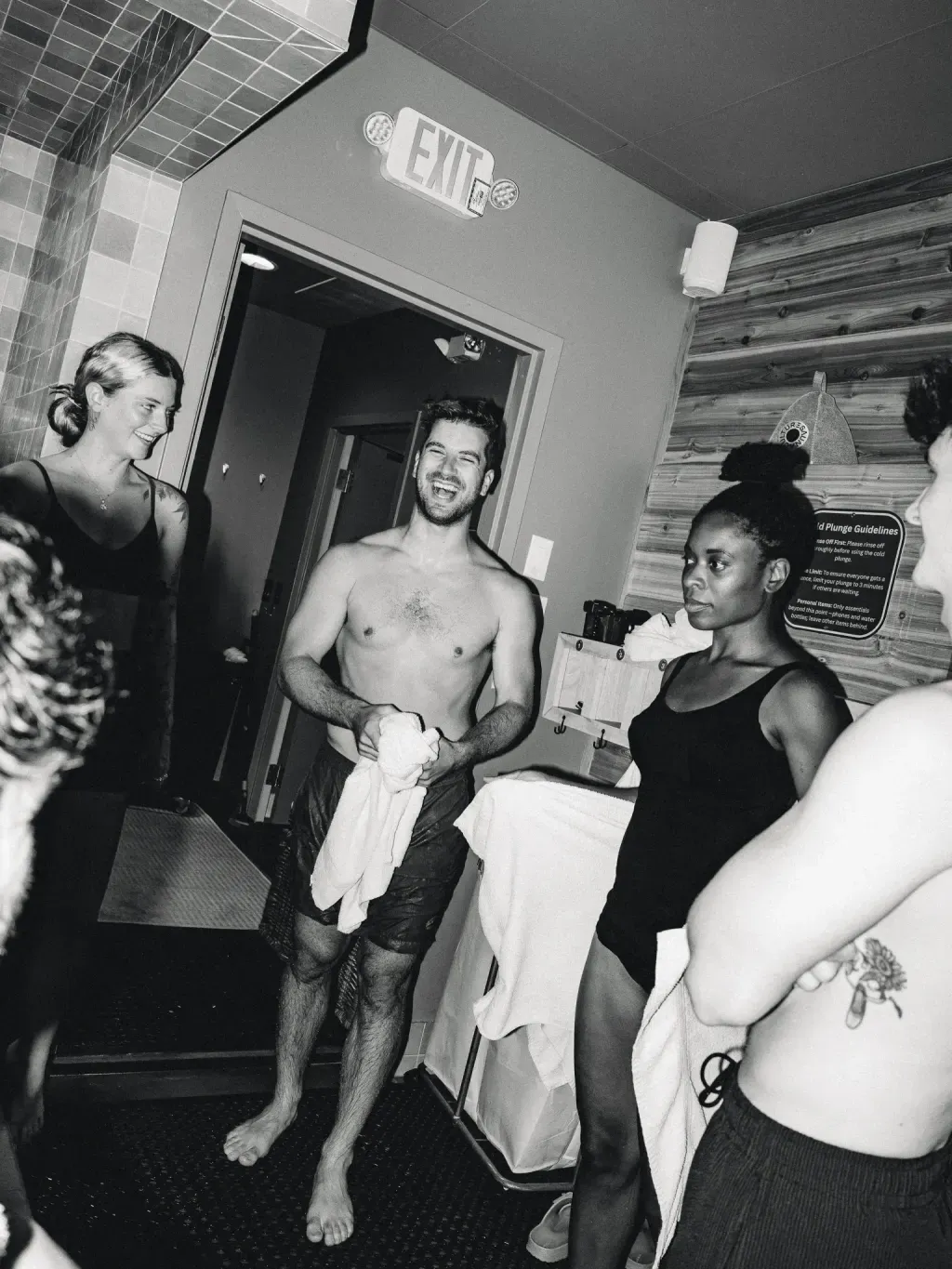 Black-and-white of friends laughing by the Cold Plunge Guidelines sign at Sauna Culture Charlotte