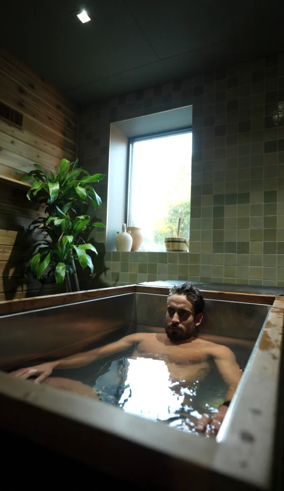 Guest in an individual cold plunge beside a window and plants at Sauna Culture Cotswold